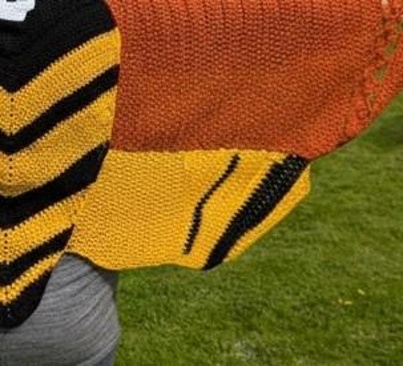 May include: A close-up of a knitted shawl with a bee wing design. The shawl features a combination of black, yellow, and orange colors. The design includes black stripes on yellow sections, and a solid orange upper portion.