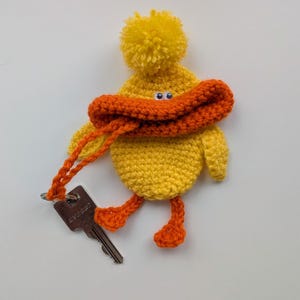 May include: A handmade yellow crochet duck keychain with orange accents. The duck has a pompom on its head, an orange beak, and orange feet. It holds a key with an orange crochet strap. The keychain is a whimsical accessory.
