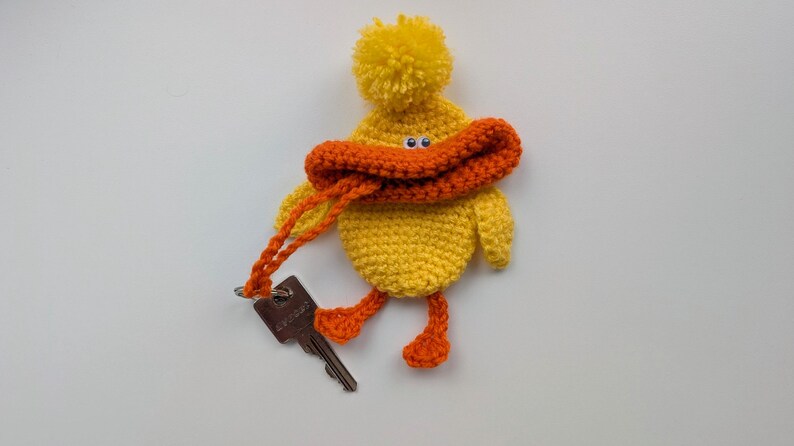 May include: A handmade yellow crochet duck keychain with orange accents. The duck has a pompom on its head, an orange beak, and orange feet. It holds a key with an orange crochet strap. The keychain is a whimsical accessory.
