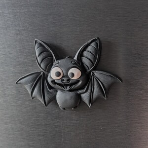 May include: A black, cartoon-style bat with large, expressive eyes and a smiling mouth. The bat has detailed wings and ears, and is set against a dark gray background. This decorative item is likely a Halloween or gothic-themed product.