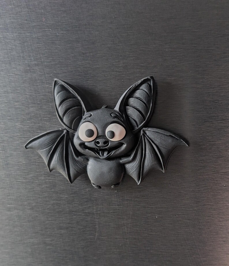 May include: A black, cartoon-style bat with large, expressive eyes and a smiling mouth. The bat has detailed wings and ears, and is set against a dark gray background. This decorative item is likely a Halloween or gothic-themed product.