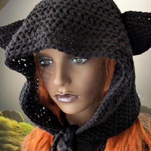 Puede incluir: Un gorro de punto negro con capucha y orejas de gato. El gorro est&aacute; hecho con un delicado patr&oacute;n de punto abierto y tiene una capucha larga y fluida.