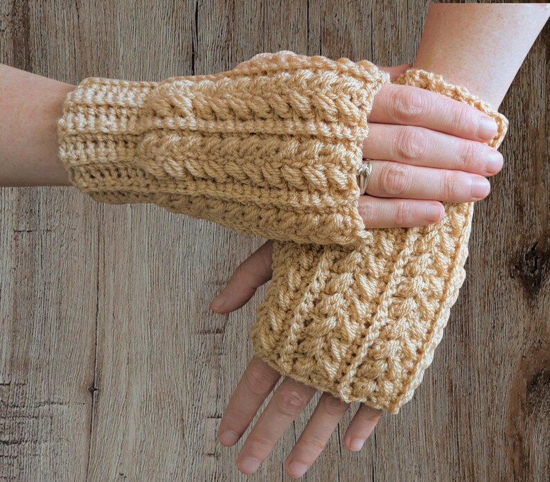 May include: A pair of light tan crochet fingerless gloves. The gloves feature a textured cable knit design. The wrist cuffs are ribbed, and the gloves cover the hand up to the knuckles, leaving the fingers free.