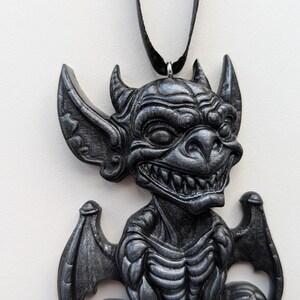 May include: A dark gray gargoyle pendant with a black cord. The pendant features a detailed, textured design of a grinning gargoyle with horns, large ears, and wings. The gothic-style necklace is a unique accessory.