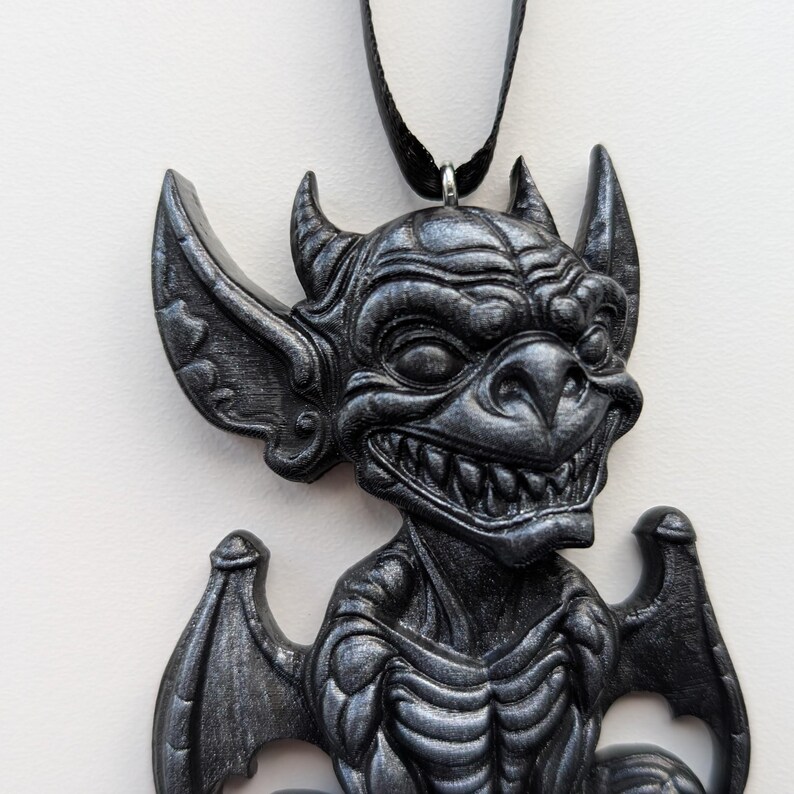 May include: A dark gray gargoyle pendant with a black cord. The pendant features a detailed, textured design of a grinning gargoyle with horns, large ears, and wings. The gothic-style necklace is a unique accessory.