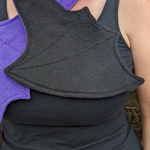 May include: A pair of purple and black fabric wings, worn on a person's shoulders. The wings are made of a soft, fuzzy material and have a pointed shape.