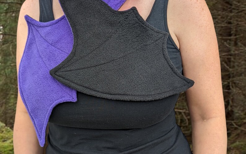 May include: A pair of purple and black fabric wings, worn on a person's shoulders. The wings are made of a soft, fuzzy material and have a pointed shape.