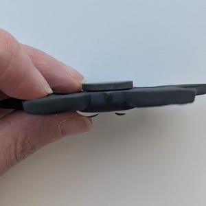 May include: A dark gray fidget spinner with a circular center and three rounded wings. The spinner has white accents and is held by a hand against a white background. The spinner is a toy.