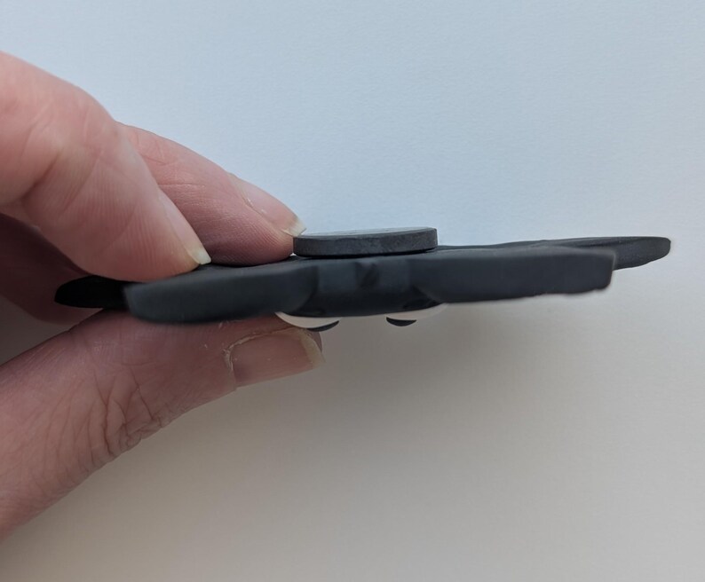 May include: A dark gray fidget spinner with a circular center and three rounded wings. The spinner has white accents and is held by a hand against a white background. The spinner is a toy.