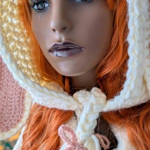 May include: A crocheted hat with a pink and white ear and a white tassel tied with a pink ribbon. The hat is made with a combination of yellow, white, and pink yarn.