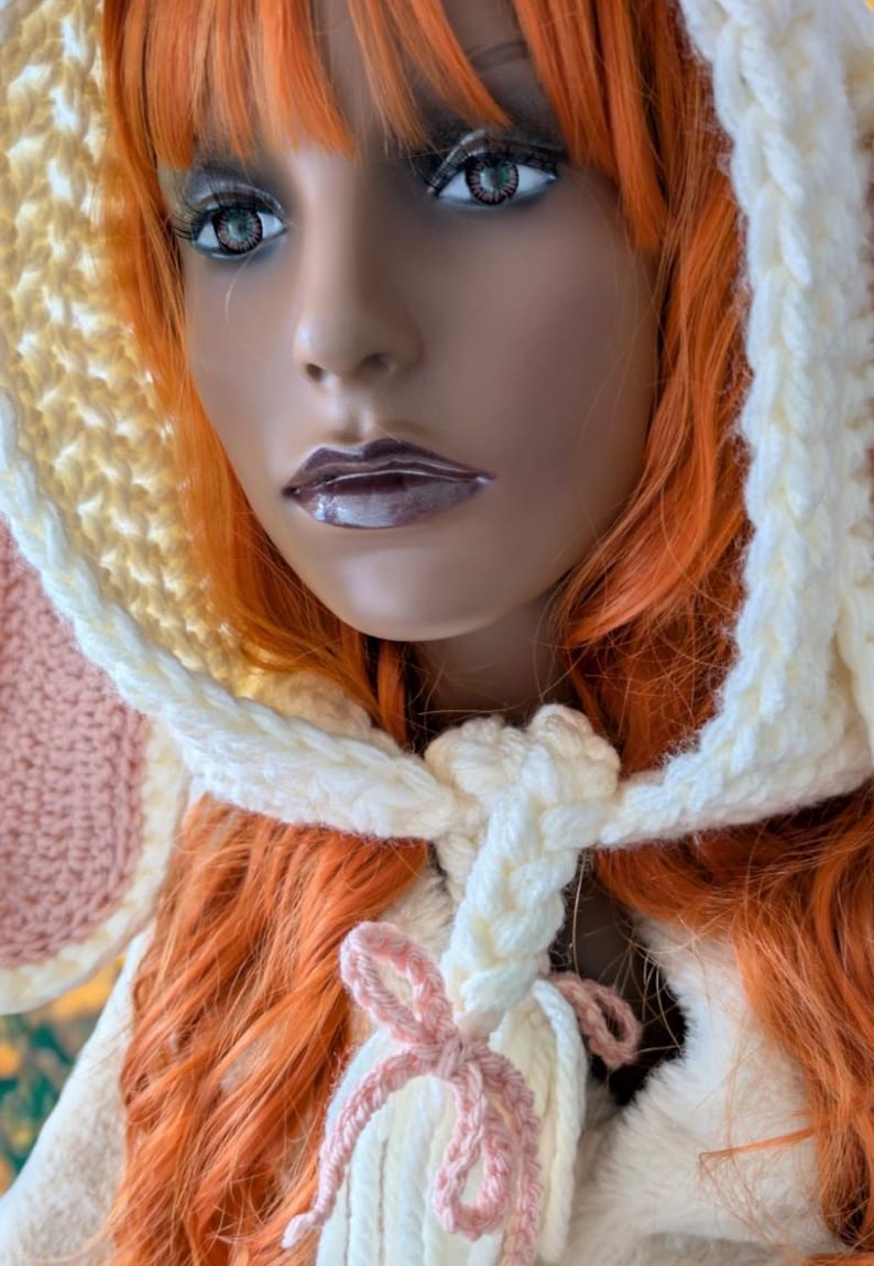 May include: A crocheted hat with a pink and white ear and a white tassel tied with a pink ribbon. The hat is made with a combination of yellow, white, and pink yarn.