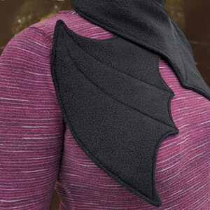 Op de afbeelding: Een zwarte fleece sjaal met een vleermuisvleugel ontwerp. De sjaal wordt om de nek gedragen en heeft een puntige kraag.
