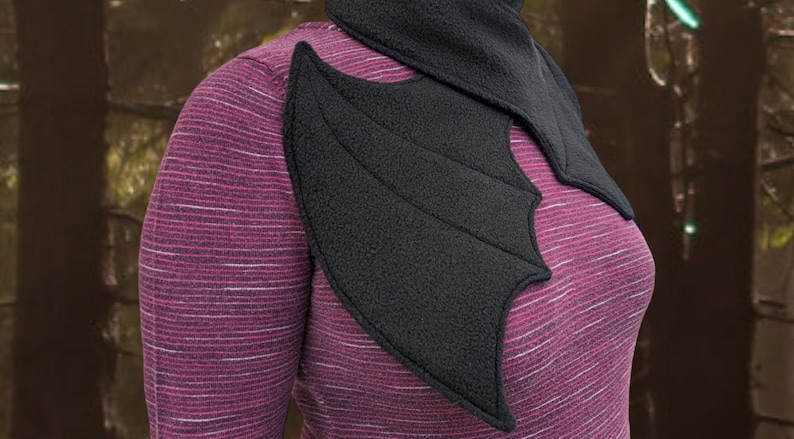 Op de afbeelding: Een zwarte fleece sjaal met een vleermuisvleugel ontwerp. De sjaal wordt om de nek gedragen en heeft een puntige kraag.