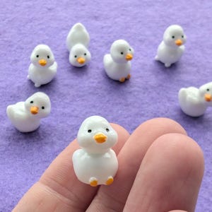 May include: A set of seven white ceramic duck figurines with orange beaks. The ducks are all in different poses, some are standing, some are sitting, and some are swimming. The ducks are all approximately 2.5 cm tall.