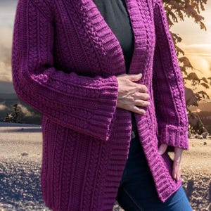 May include: A long, open-front, purple crochet cardigan with a textured pattern. The sleeves are long and cuffed. The cardigan is worn over a black top and blue jeans. The background shows a snowy landscape with a sunset.