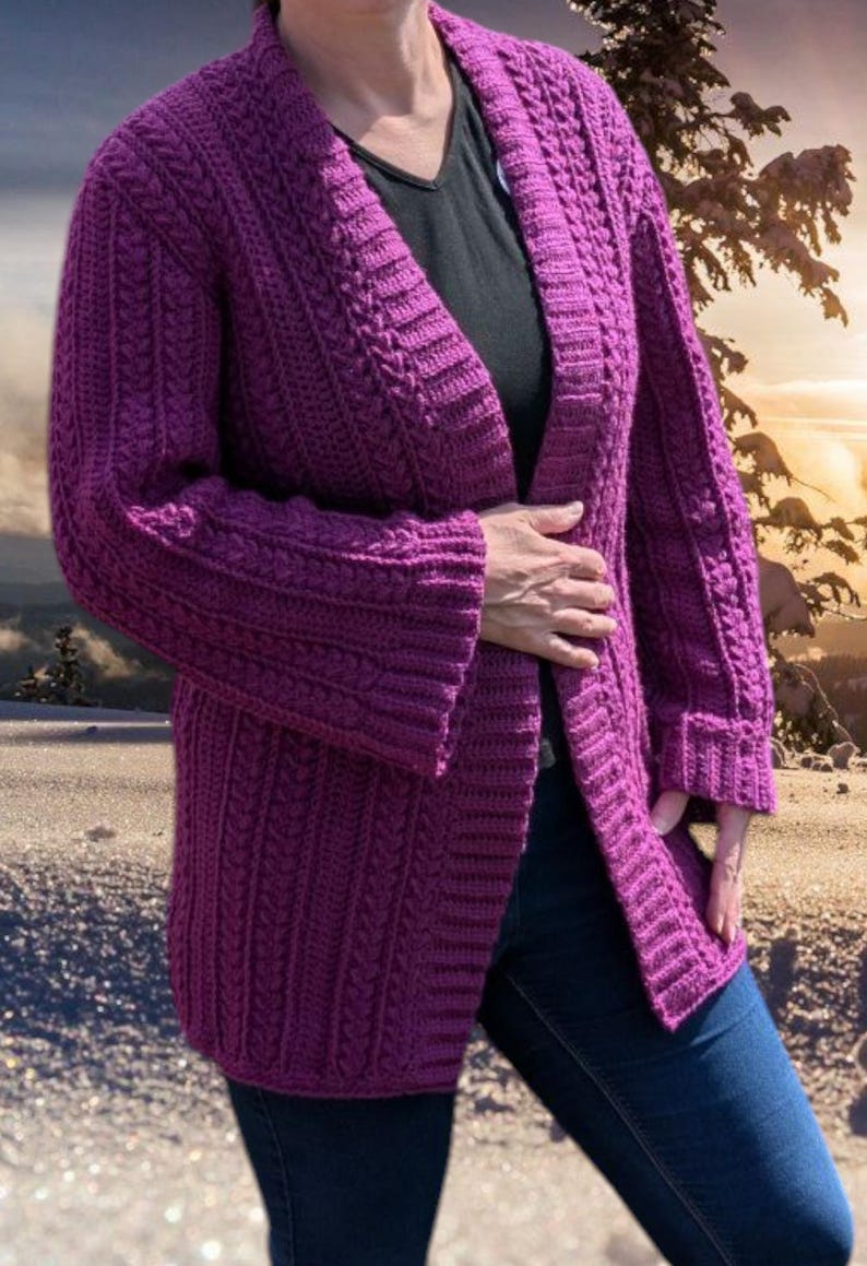 May include: A long, open-front, purple crochet cardigan with a textured pattern. The sleeves are long and cuffed. The cardigan is worn over a black top and blue jeans. The background shows a snowy landscape with a sunset.
