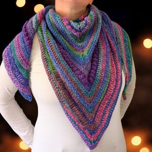 Pu&ograve; includere: Uno scialle all'uncinetto fatto a mano, di forma triangolare e multicolore. Lo scialle presenta sfumature di viola, blu, verde e rosa. Il filato &egrave; strutturato e lo scialle si indossa intorno al collo e alle spalle.