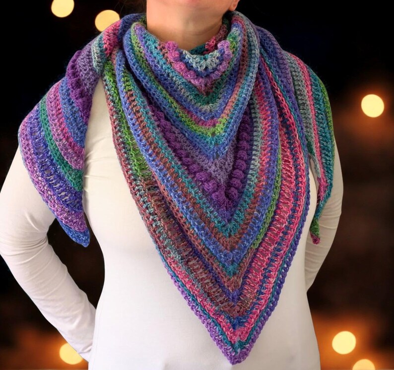 Pu&ograve; includere: Uno scialle all'uncinetto fatto a mano, di forma triangolare e multicolore. Lo scialle presenta sfumature di viola, blu, verde e rosa. Il filato &egrave; strutturato e lo scialle si indossa intorno al collo e alle spalle.