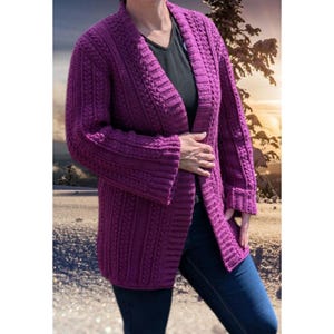 May include: A long, open-front cardigan in a deep purple hue. The knitted sweater features a textured cable knit design on the sleeves and front panels. The cardigan is worn over a black top and blue jeans.