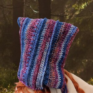 May include: A crocheted hooded cowl in shades of purple, blue, and pink. The cowl is made with a striped pattern and has a long, flowing design.