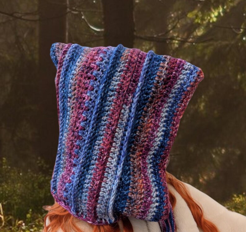 May include: A crocheted hooded cowl in shades of purple, blue, and pink. The cowl is made with a striped pattern and has a long, flowing design.