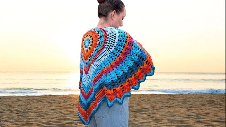 May include: A colorful, crocheted shawl in shades of blue, orange, and pink. The shawl features a circular design on the back and a scalloped edge. The person is standing on a sandy beach with the ocean in the background.