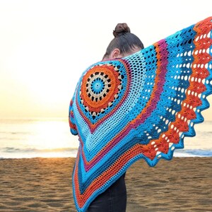 May include: A colorful, handmade crochet shawl with a circular design in shades of blue, orange, and pink. The shawl is being held up on a beach with the ocean and sunset in the background. The shawl's dimensions are approximately 60 inches wide.