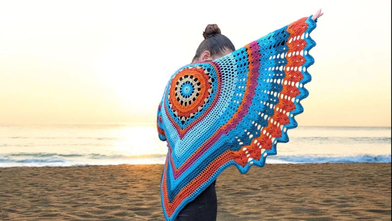 Op de afbeelding: Een levendige, handgemaakte gehaakte sjaal in tinten blauw, oranje en roze. De sjaal heeft een cirkelvormig ontwerp in het midden en een geschulpte rand. De persoon staat op een strand met de oceaan en zonsondergang op de achtergrond.