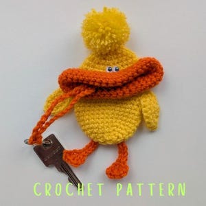 May include: A handmade yellow crochet duck keychain with an orange beak, feet, and a pom-pom hat. The duck has googly eyes and is holding a key. The text "CROCHET PATTERN" is at the bottom.