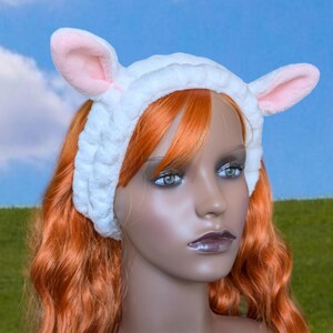 May include: A white terry cloth headband with two pink-lined ears. The headband is designed to resemble a sheep. The model in the image has orange hair and is wearing the headband.