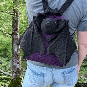 May include: A handmade crochet backpack shaped like a bat. The backpack features black wings, a black head with red eyes, and a purple interior. The straps and back of the bag are black.