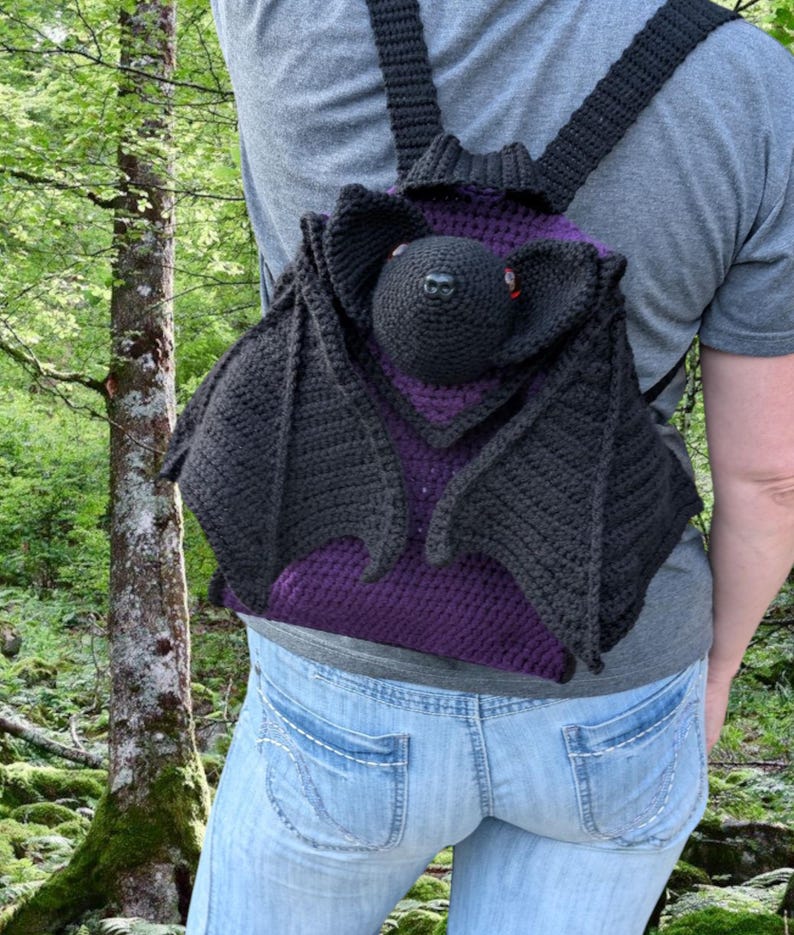 May include: A handmade crochet backpack shaped like a bat. The backpack features black wings, a black head with red eyes, and a purple interior. The straps and back of the bag are black.