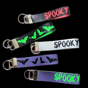 May include: A collection of Halloween-themed keychains. The keychains feature various designs, including the word "Spooky" in dripping font and bat silhouettes. The keychains are in black, silver, and purple with neon green and red accents.