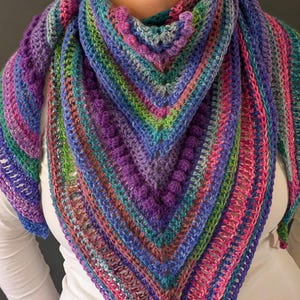 Pu&ograve; includere: Uno scialle all'uncinetto fatto a mano e colorato a forma triangolare. Lo scialle presenta un vivace mix di colori, tra cui viola, blu, verde e rosa. Il design incorpora punti strutturati e un motivo a righe, creando un accessorio accogliente ed elegante.