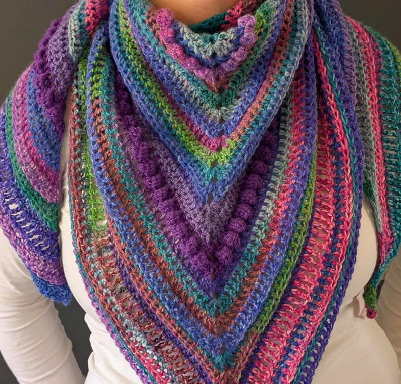 Pu&ograve; includere: Uno scialle all'uncinetto fatto a mano e colorato a forma triangolare. Lo scialle presenta un vivace mix di colori, tra cui viola, blu, verde e rosa. Il design incorpora punti strutturati e un motivo a righe, creando un accessorio accogliente ed elegante.