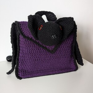 May include: A handmade crochet bag in the shape of a bat. The bag is primarily purple with black trim and a black bat head with red eyes. The bag has a handle and shoulder straps.