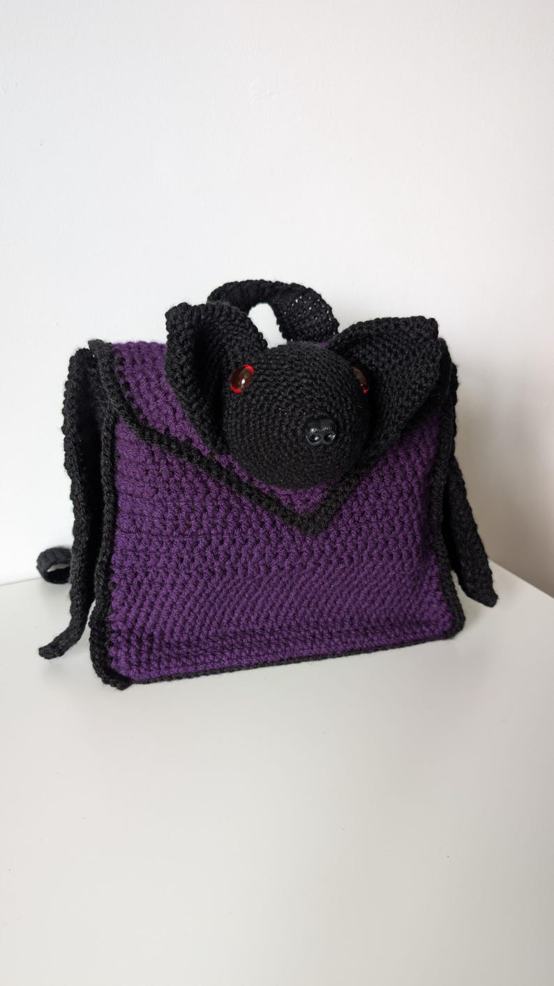 May include: A handmade crochet bag in the shape of a bat. The bag is primarily purple with black trim and a black bat head with red eyes. The bag has a handle and shoulder straps.