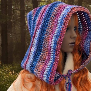 May include: A colorful crocheted hooded scarf with a striped pattern in shades of blue, pink, and purple. The scarf is worn by a person with long, red hair.