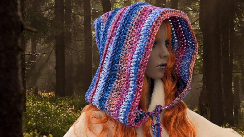 May include: A colorful crocheted hooded scarf with a striped pattern in shades of blue, pink, and purple. The scarf is worn by a person with long, red hair.