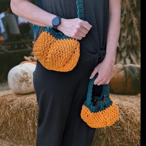 Op de afbeelding: Twee gehaakte tassen, één als crossbody en de andere in de hand. De tassen zijn pompoenkleurig met teal accenten. De tassen zijn gemaakt van een dik, getextureerd garen.