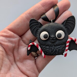 Puede incluir: Adorno de murci&eacute;lago negro con grandes ojos blancos y alas de bast&oacute;n de caramelo. El murci&eacute;lago tiene un pecho peludo y orejas grandes. Se adjunta una cinta negra para colgar. Ideal para Halloween o Navidad.