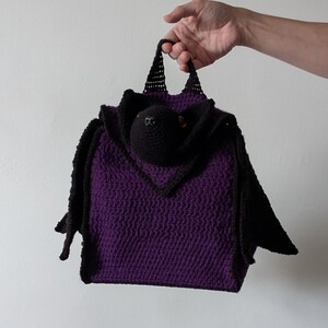 May include: A handmade purple crochet bag with a black bat design. The bag features a bat head with red eyes and black wings. The bag is held by a hand against a white background.