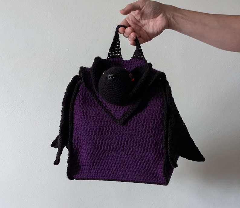 May include: A handmade purple crochet bag with a black bat design. The bag features a bat head with red eyes and black wings. The bag is held by a hand against a white background.