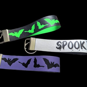 May include: Three Halloween-themed keychains with silver hardware. One is black with green bats, another is silver with the word "SPOOKY" in dripping black letters, and the third is purple with black bats.