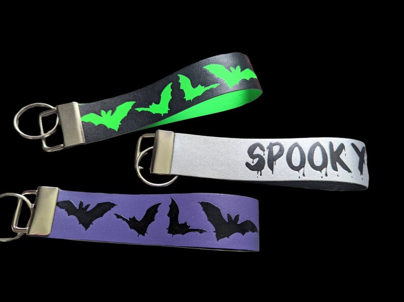 May include: Three Halloween-themed keychains with silver hardware. One is black with green bats, another is silver with the word "SPOOKY" in dripping black letters, and the third is purple with black bats.
