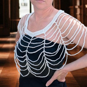 May include: A white, open-weave crochet top with a unique web-like design. The top features a scoop neckline and delicate, dangling strands, creating a stylish, airy look. The garment is worn over a black top, offering a striking contrast.