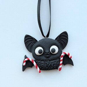 Puede incluir: Adorno de murci&eacute;lago negro con ojos blancos grandes y detalles de bastones de caramelo. El murci&eacute;lago tiene orejas grandes y cuelga de una cinta negra. Una decoraci&oacute;n festiva para Halloween o Navidad.