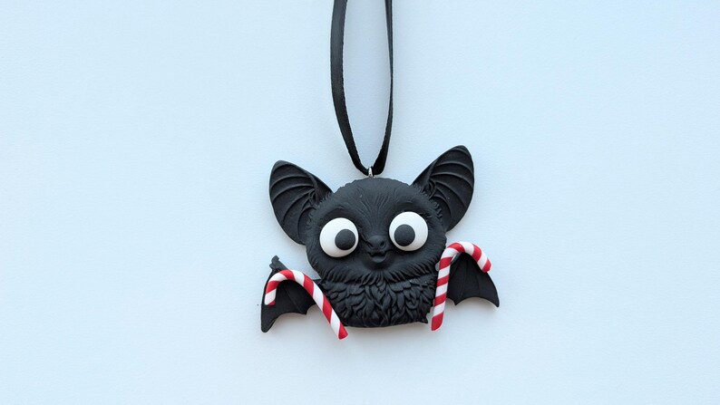 Puede incluir: Adorno de murci&eacute;lago negro con ojos blancos grandes y detalles de bastones de caramelo. El murci&eacute;lago tiene orejas grandes y cuelga de una cinta negra. Una decoraci&oacute;n festiva para Halloween o Navidad.