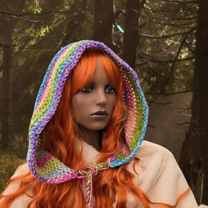 May include: A woman with long, bright orange hair wears a colourful crocheted hooded scarf. The scarf is made with a variety of colours, including purple, blue, green, and pink. The woman is wearing a white hooded robe.