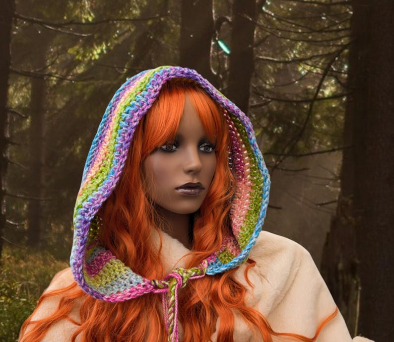 May include: A woman with long, bright orange hair wears a colourful crocheted hooded scarf. The scarf is made with a variety of colours, including purple, blue, green, and pink. The woman is wearing a white hooded robe.